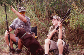 Photo of man, boy, and dog hunting in field.