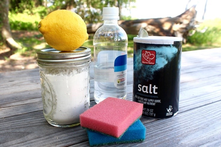different cleaning products including; a lemon, water, salt, detergent, and sponges. All sitting on a wooden table outdoors.
