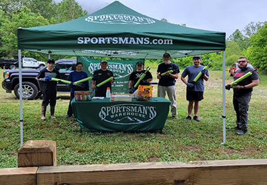 A group of people standing under a Sportsman's Warehouse tent, holding clean-up gear.