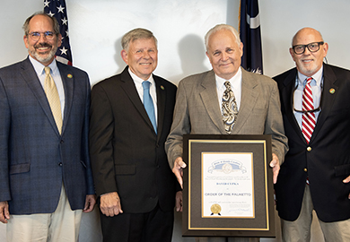 Four people in suits smiling at the camera. One of them, David Cupka, in the middle is holding a framed award.