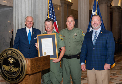 Two men in suits and two men in uniform standing together. One uniformed man holds a framed document.