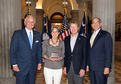 Woman holding award standing next to three men. United States and South Carolina flags in the background.