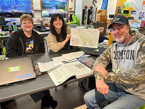 Two women and a man sitting at a classroom table. One of the women is holding a drawing of a bird